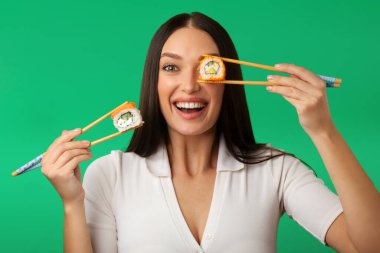 İki elinde yemek çubuklarıyla suşi tutan duygusal genç bir kadın, bir gözü kaplayan, yeşil arka planda duran.