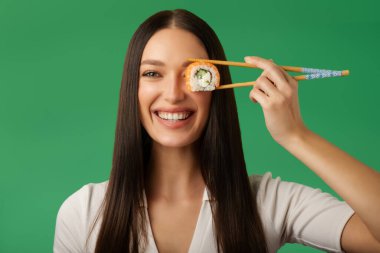 Suşi rulosundan hoşlanan mutlu bir bayan, onu yemek çubuklarıyla gözünün önünde tutuyor ve gülümsüyor, yeşil arka planda poz veriyor.