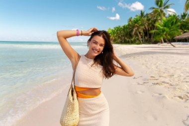 Kumsalda gülümseyen genç bir kadın, tropik bir adada tatilin tadını çıkarıyor, arka planda palmiyeler.