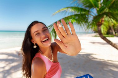 Güneş kremi şişesini tutan genç ve mutlu bir bayan. Plajda durup kameraya gülümsüyor.