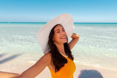 Sarı mayo ve hasır şapkalı güzel mutlu kadın okyanus kıyısında duruyor ve gülümsüyor, tropikal havanın ve güneşin tadını çıkarıyor.