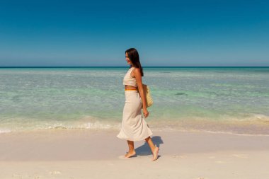 Çantalı kadın turist tropikal adada yürüyor, turkuaz okyanus suyunun ve kumlu kumsalın tadını çıkarıyor.