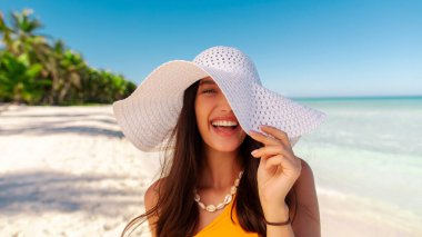 Sahilde arka planda açık mavi okyanusla duran hasır şapkalı genç kadın portresi, kameraya gülümseyen kadın, kumlu sahilde yürüyen kadın.