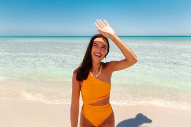 Kumsalda gülümseyen mutlu genç bir kadın, kendini güneşten koruyor, arka plandaki turkuaz okyanusun cennet adasında tatilin tadını çıkarıyor.