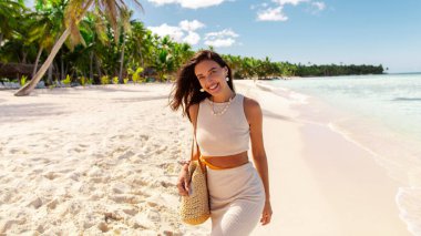 Kumsalda gülümseyen genç bir kadın, tropik bir adada tatilin tadını çıkarıyor, arka planda palmiyeler.