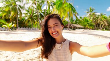 Mutlu gülümseyen genç bir kadın sahilde selfie çekiyor, tropik bir adada tatilin tadını çıkarıyor, arka planda avuçlar.