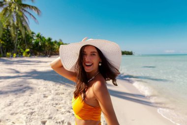 Sahilde hasır şapka takan, arka planında turkuaz okyanus olan güzel, mutlu bir bayan kameraya bakıyor ve gülüyor, tropikal tatilin tadını çıkarıyor.