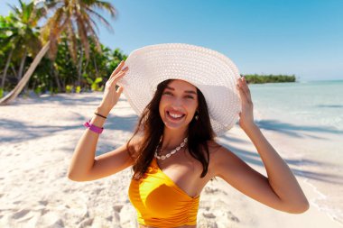 Sahilde hasır şapka takan, arka planında turkuaz okyanus olan güzel, mutlu bir bayan kameraya bakıyor ve gülüyor, tropikal tatilin tadını çıkarıyor.