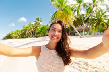 Mutlu gülümseyen genç bir kadın sahilde selfie çekiyor, tropik bir adada tatilin tadını çıkarıyor, arka planda avuçlar.