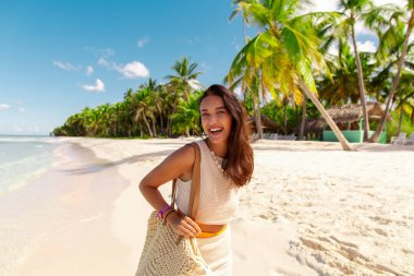 Kumsalda gülümseyen genç bir kadın, tropik bir adada tatilin tadını çıkarıyor, arka planda palmiyeler.