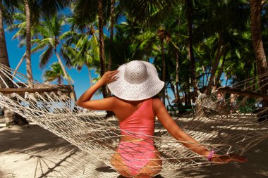 Hasır şapkalı, tanınmayan genç bir kadın cennetteki bir plajda, palmiye ağaçlarının altında oturuyor.