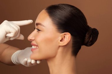 Estetik ameliyat ve güzellik konsepti. Kadın burnuna dokunan bir cerrah. Kahverengi arka plandaki burun ameliyatından önce danışmanlık yapıyor.