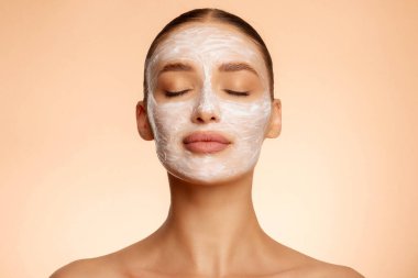 Besleyici bir yüz bakımı. Güzellik ameliyatı yapan kadın yüz kilinden maske takıyor, bej arka planda, gözleri kapalı poz veriyor.