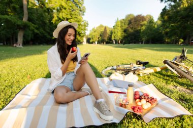 Mutlu bayan güneşli parkta piknik yapıyor, battaniyeye oturmuş taze meyve yerken cep telefonunu kaydırıyor.