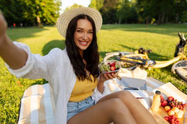 Sunny Park 'ta piknik battaniyesine oturmuş elinde taze meyve tabağıyla akıllı telefondan neşeli bir selfie çeken ve kameraya gülümseyen genç bir kadın.