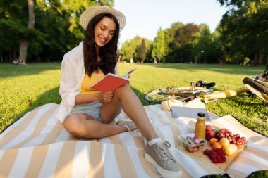 Güneşli parkta battaniyeye oturmuş dışarıda eğlenirken kitap okuyor ve yalnız piknik yapıyor. Temiz hava çıkışı.