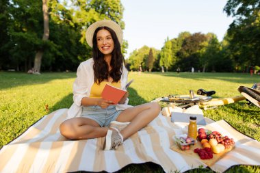 Güneşli boş zaman. Mutlu kadın parkta piknik battaniyesinde oturmuş kitap okuyor ve taze meyvelerin tadını çıkarıyor.