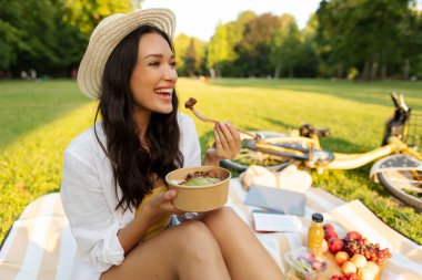 Taze yiyecek getirdim. Kadın kaseden yemek yiyor, güneşli parkın rahatlatıcı atmosferinde battaniyeye oturuyor.