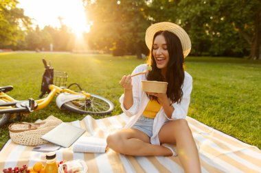 Kadın battaniyeye oturmuş, güneşli yaz parkında piknik yaparken kaseden taze salata yer gibi gülümsüyor. Meyveler ve ekose üzerine açık bir kitap.
