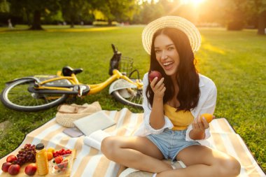 Yaz pikniği eğlencesi. Güneşli yaz parkında battaniyeye oturmuş kameraya bakarken elinde taze meyve tutan mutlu kadın.