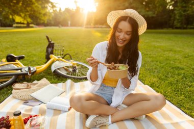 Parkta piknik battaniyesinde oturan ve kaseden salata yerken gülümseyen, güneşli yaz gününün tadını çıkaran, yakınlardaki çimlerin üzerinde uzanan bisikletli bir kadın.