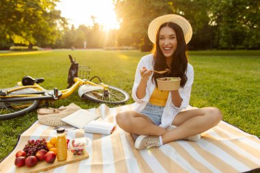 Şehirdeki sağlıklı öğle yemeğini seven bir kadın, parkta piknik yaparken battaniyeye oturmuş, yakınlardaki çimlerin üzerinde yatan bir bisiklet.