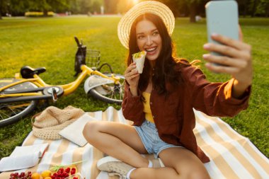 Temiz hava yemeği. Mutlu bayan cep telefonuyla selfie çekerken abur cubur yiyor, piknik yapıyor, güneşli bir günde dışarıda takılıyor.