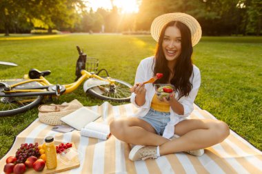 Taze yaz atıştırması. Meyve yiyen ve kameraya gülümseyen mutlu kadın, sıcak güneş ışığıyla parkta battaniyeye oturmuş, bisiklet ona yakın.