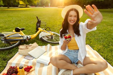Yeşil çimenlerin üzerinde piknik battaniyesinde oturan gülümseyen kadın elinde taze meyve kasesi ve sıcak yaz güneşinde mutlu bir şekilde kameraya bakıyor. Canlı piknik sahnesi