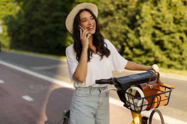 Güneşli bir günde parkta elektrikli bisikletin yanında dikilirken cep telefonuyla konuşan mutlu bir bayan modern ulaşımı kullanarak özel bir yolda bisiklet sürüyor.
