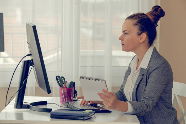 Side view of a girl in white shirt and plaid jacket near her computer in the office. She is looking at the screen and holding a notebook. Online working and education.. Concept of office routine.