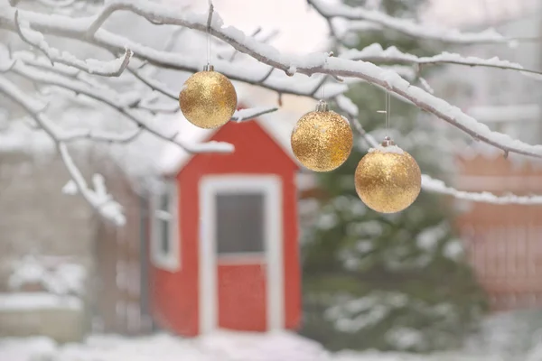 Χρυσές μπάλες χριστουγεννιάτικου δέντρου στα κλαδιά και το χιόνι του χειμώνα. Χριστουγεννιάτικες μπάλες κοντά σε τριανταφυλλιές καλυμμένες με χιόνι και μικρό ξύλινο σπίτι. — Φωτογραφία Αρχείου