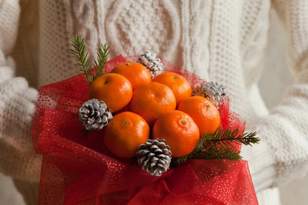 Woman hands in white sweater holding bouquet of mandarins and Christmas tree branches. New Years edible bouquet of fruits. Gift for Christmas. DIY gift. useful gift made of fruits. decor of fruits.