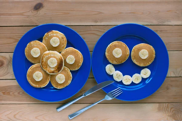 Pancakes with smile on blue plate and wooden background. banana fruit smiling breakfast - fun food for kids — Stock Photo, Image