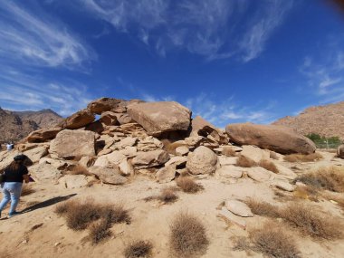 Tabuk 'taki Altın Kaya bölgesinin çarpıcı gündüz perspektifi. Tarihi kırmızı kayalar, parlayan çöl kumları ve berrak mavi gökyüzü birleşerek pitoresk bir manzara oluşturuyor..