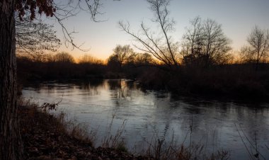 Sakin bir nehir, alacakaranlıkta sessiz bir ormanlık arazide yavaşça akar, batan güneşin sıcak, huzurlu parıltısıyla yıkanır, sakin ve doğal güzelliği anımsatır..