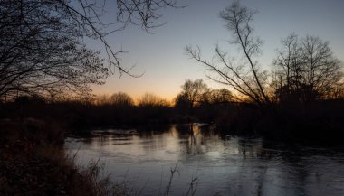 Sakin bir akşam sahnesi, sakin suya yansıyan alacakaranlığın yumuşak parıltısını yakalar, ağaçların ve sazlıkların karanlık siluetleriyle çevrili, derin bir huzur hissi uyandırır..