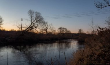 Sakin bir nehir, alacakaranlıkta sessiz ve çıplak bir ormanın içinden akar. Yumuşak, berrak alacakaranlık gökyüzünü yansıtır ve huzur dolu bir yalnızlık hissi uyandırır..