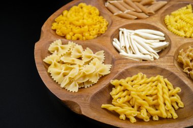 Various mix of pasta on wooden bowl and black background.