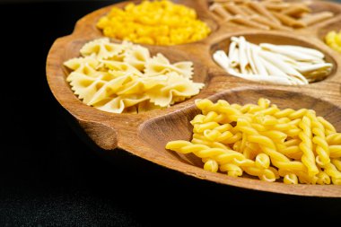 Various mix of pasta on wooden bowl and black background.