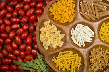 Various mix of pasta on wooden bowl, small red tomatoes on black