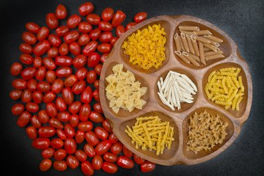 Various mix of pasta on wooden bowl, small red tomatoes on black