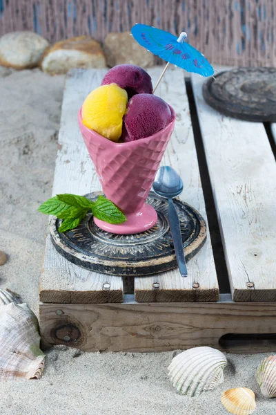 Mango and black-currant ice-cream scoops in pink cone on the beach — Stock Photo, Image