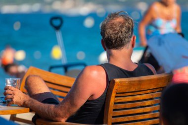 Yazın Fransa 'da Saint-Tropez Körfezi' nin mavi sularına bakan kimliği belirsiz bir adam bir bardak içkiyle sahil kafesinde oturuyor.