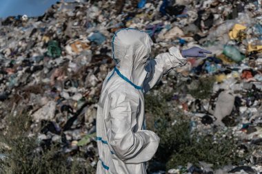 Individual in protective suit assesses a massive pile of rubbish at a landfill site, highlighting the issue of waste management and pollution.