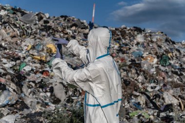 A person in protective gear uses a device to observe the landfill, surrounded by heaps of garbage and various types of pollution.