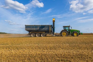 Bir traktör, hasat edilmiş bir tarladan tahıl arabasını çekiyor, açık mavi gökyüzü ve kabarık bulutların altında mahsul topluyor, tarımsal çalışmalar sergiliyor..