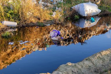 Kirli suyla dolu ve etrafı çöplerle çevrili bir kanal, bölgedeki ciddi kirlilik ve ekolojik kaygıları vurguluyor..
