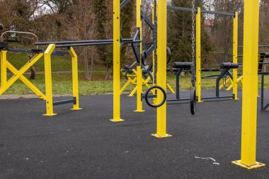 İnsanlar, sonbaharda bulutlu bir günde ağaçlı bir parkta bulunan açık hava spor salonundaki sarı egzersiz istasyonlarını kullanabilirler..