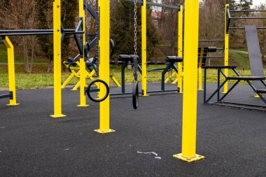 Sahne, parkta sarı egzersiz ekipmanlarının olduğu bir çalışma alanını gösteriyor. Zemin siyah kauçuk ve arka planda ağaçlar var..